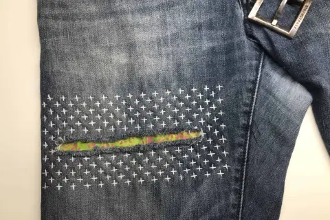 Close-up of denim jeans with a colorful fabric patch and white cross-stitch embroidery, showcasing a stylish sashiko jeans repair.