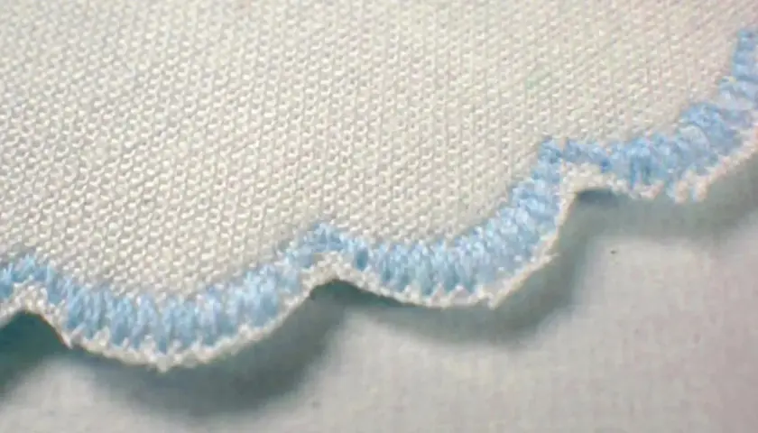 Close-up of a white fabric with a scalloped edge finished with light blue stitching. This decorative edge is called a picot.