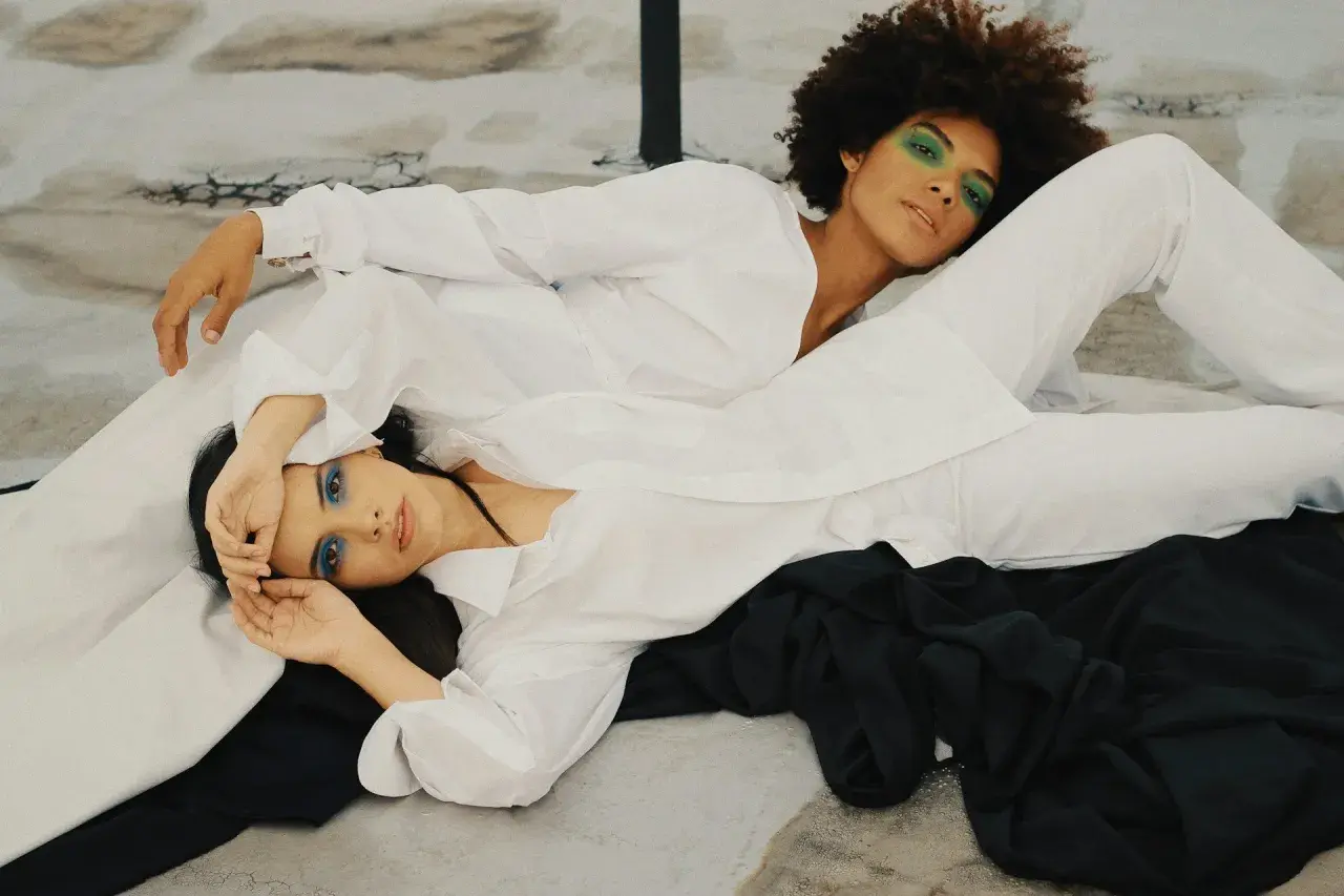 Two women in white shirts and pants, one with green eyeshadow, the other with blue. The fabric looks smooth, suggesting lyocell does not wrinkle easily.