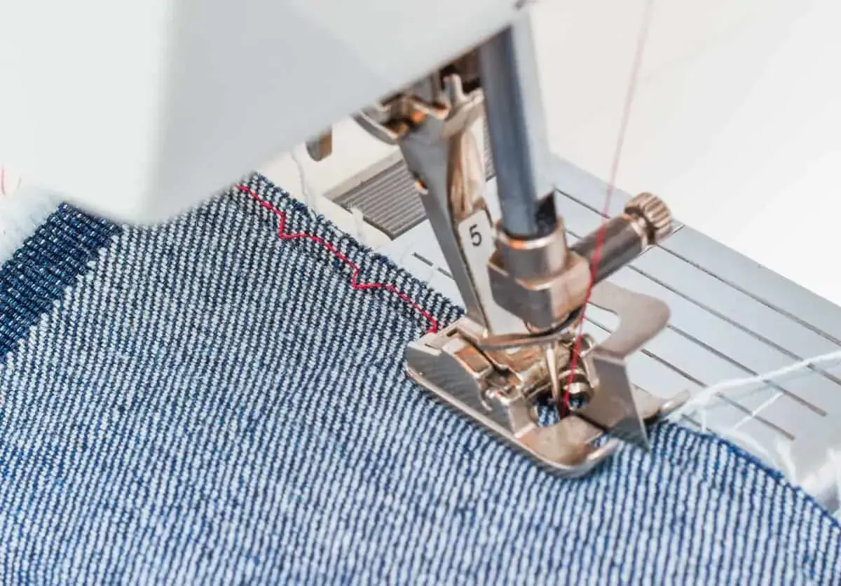 Sewing machine foot guiding denim fabric, demonstrating how to blind hem pants with a red thread stitch.