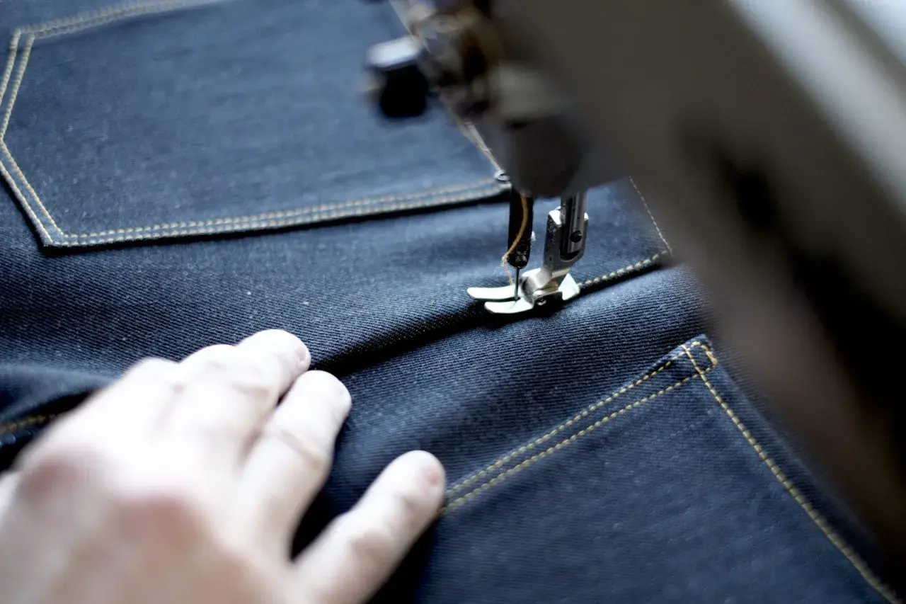 A sewing machine stitches a precise jean seam on dark denim, with a hand guiding the fabric.