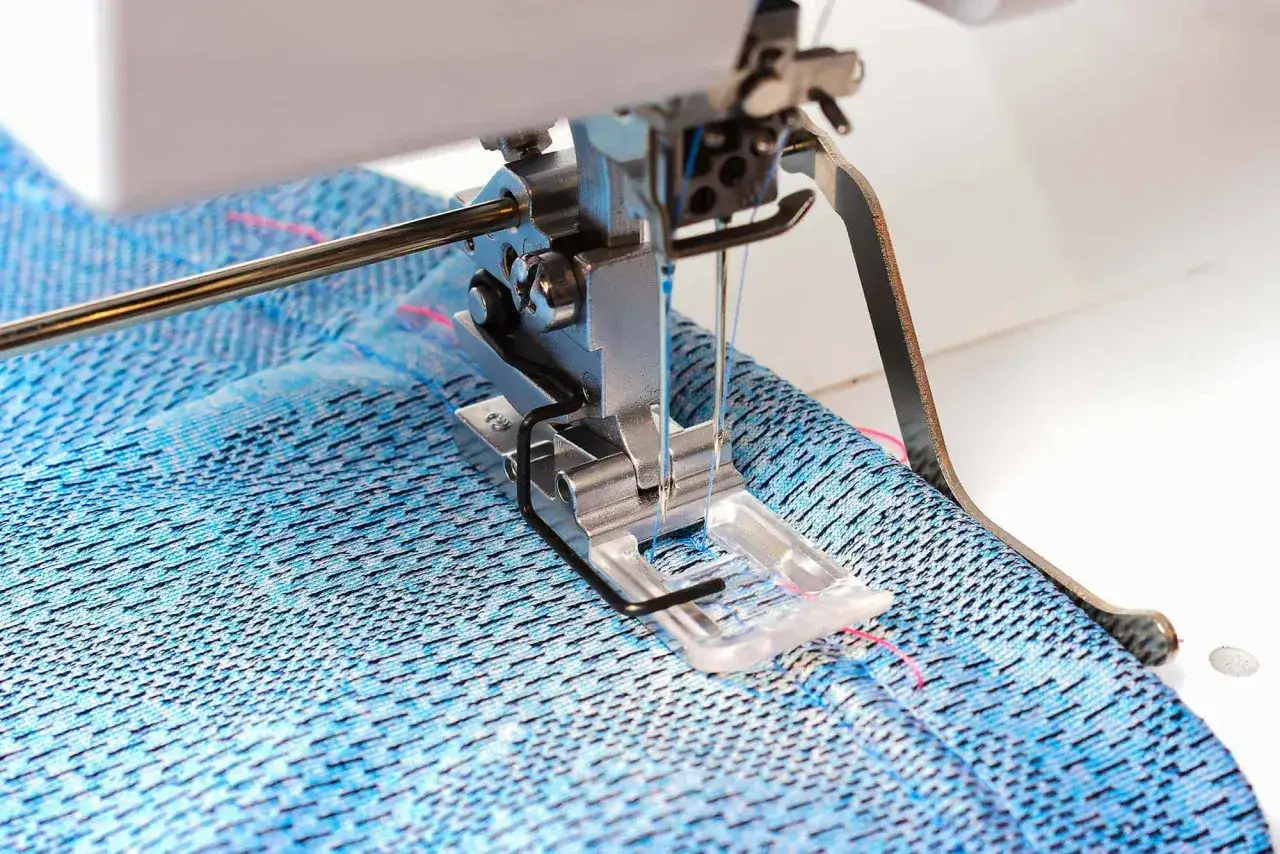 A sewing machine's presser foot guides blue fabric as it creates a coverstitch hem.