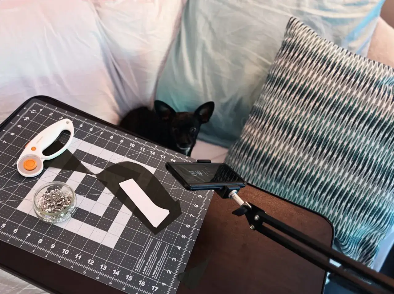 A small black dog watches as someone uses adaptive sewing tools, including a rotary cutter and pins, on a cutting mat.