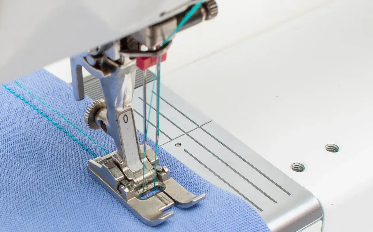 A sewing machine's double needle stitches parallel lines of turquoise thread on blue fabric. This demonstrates how a double needle can be used on many sewing machines.