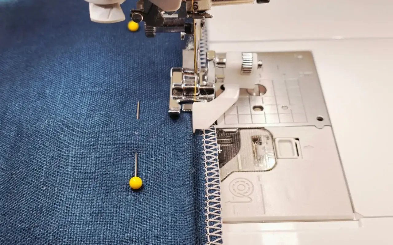 Close-up of a hemming sewing machine creating a decorative stitch on blue fabric. Pins hold the fabric in place.